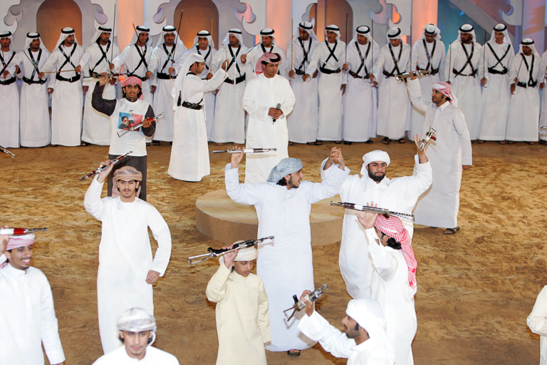 Young men hold toy guns in the air and dance in a sand-filled room lined with standing men.