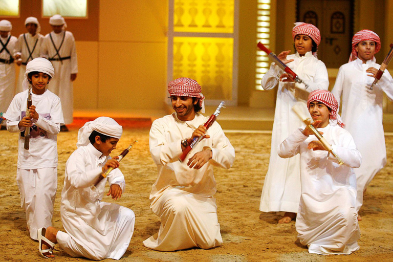 Five young boys in white robes and red checked head coverings hold toy guns and gather around a young man kneeling in sand.