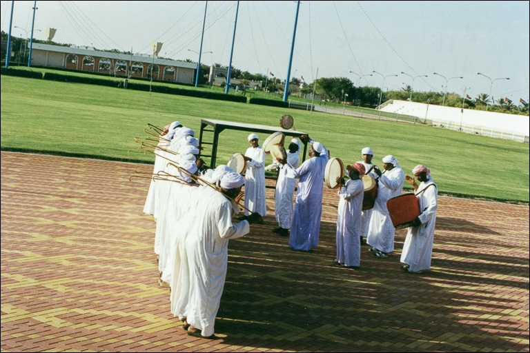 Men in white robes stand in a row near an athletic field facing other men holding instruments.