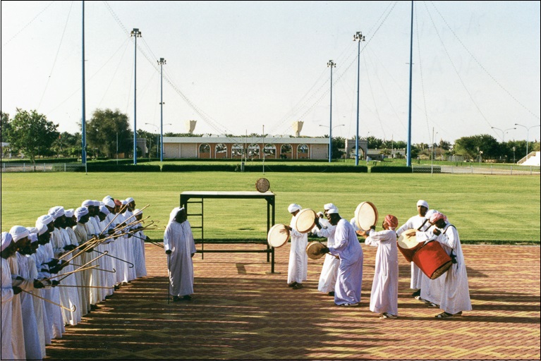 Men in white robes stand in a row near an athletic field and face other men who are playing instruments.
