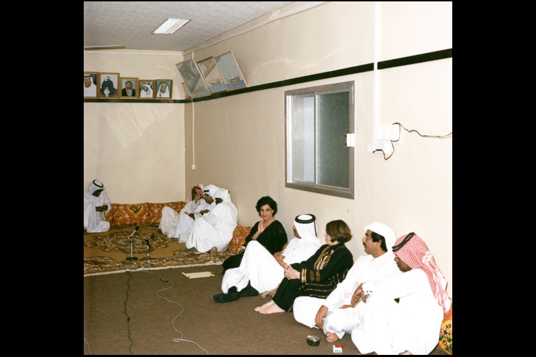 Some men and women sit on the floor and lean against a wall and a some men sit eleswhere in the room around a microphone placed on a rug. 