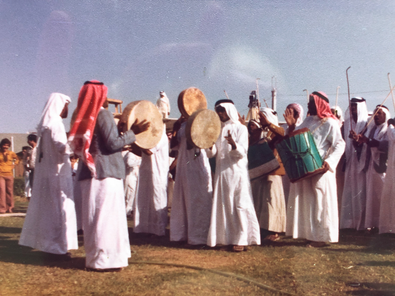 Men in white robes and head coverings perform on their drums in an outdoor setting while others watch.