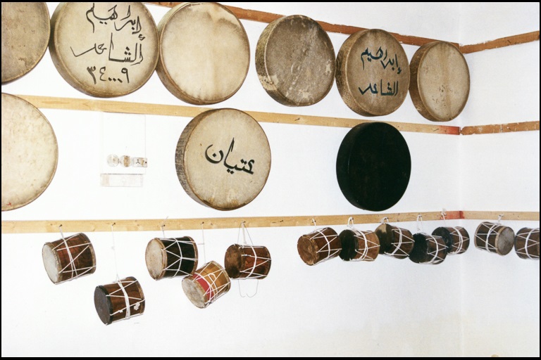 Various hand drums, some written with arabic script, hang on a white wall.