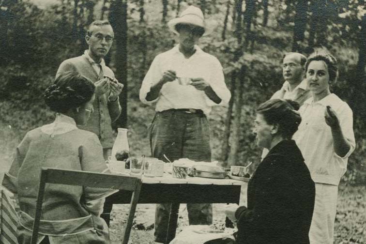 Black and white image of three women and three men eating outdoors at a camp table with woods in the background. Two of the women are seated on camp chairs.