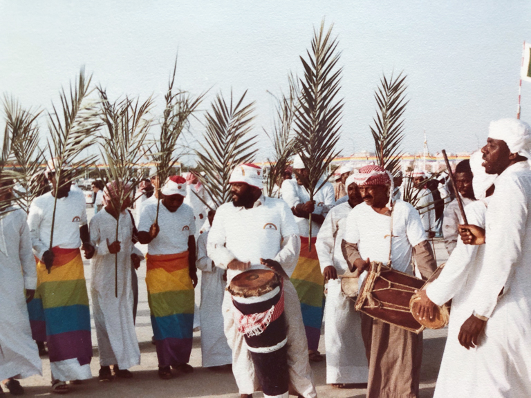 A group male drummers and singers perform and wave palm branches outdoors.