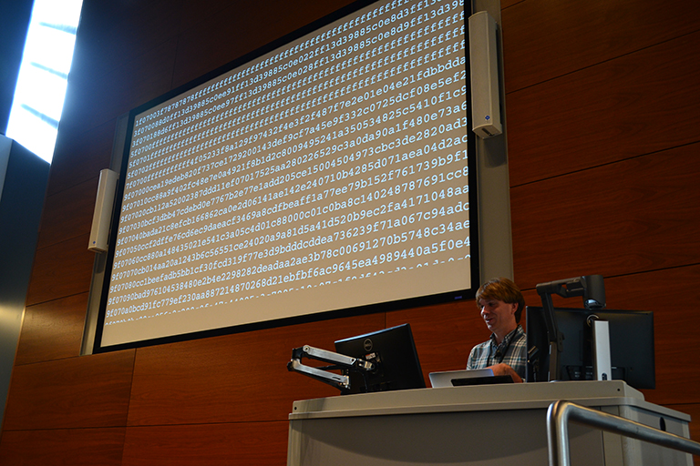 A women is presenting at a podium with a large screen above her filled with binary numbers.