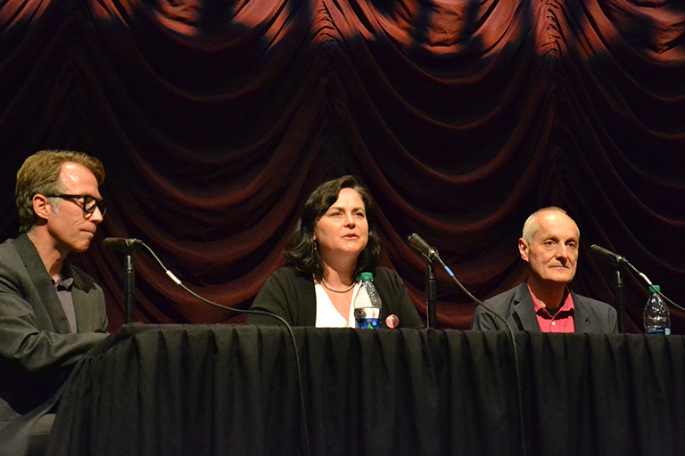 A group of three people sit in the front of a theater in a panel discussion.