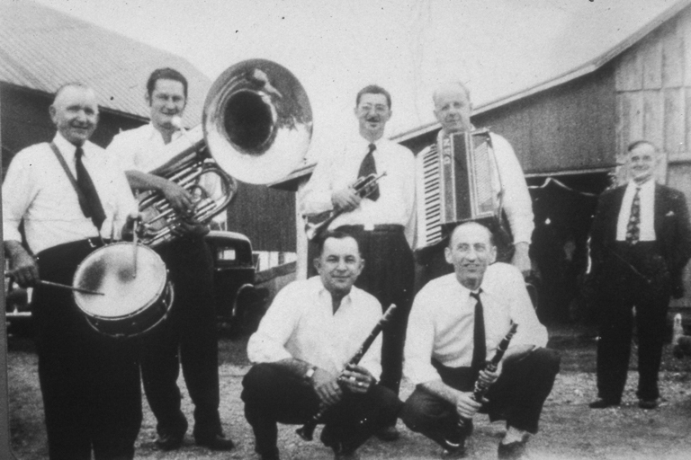 A polka band of six men poses in front of a barn.