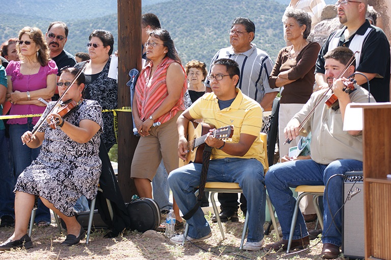 People stand behind three musicians playing fiddles and a guitar. They all look forward and are surrounded by desert mountains.