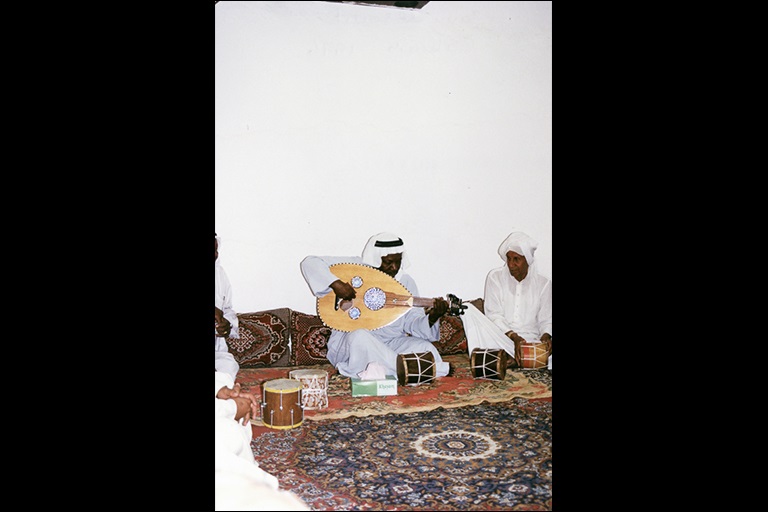 A man sitting on pillows and an oriental rug plays an ‘ūd while other men watch.