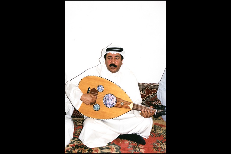 A man sits on a oriental rug and plays an ‘ūd.