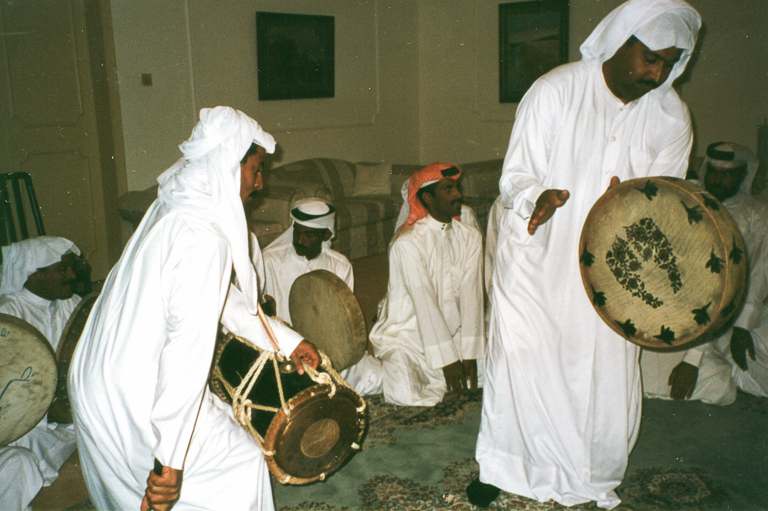 Men in white robes sit on the floor and play instruments while two other men stand in the center and play drums.