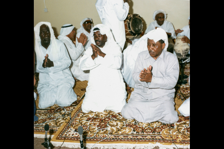 Several men in white robes kneel on an oriental rug and clap their hands.