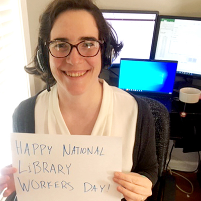 Smiling woman with glasses holding placard that reads happy national library workers day!