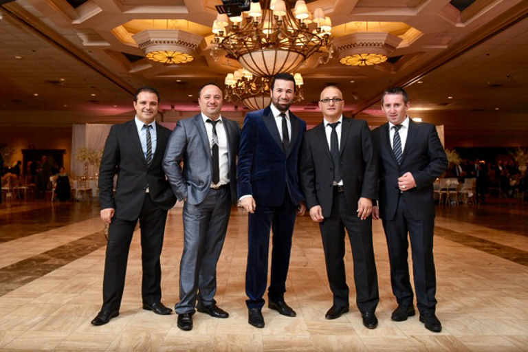 Five men in suits stand in a fancy ball room facing the camera.