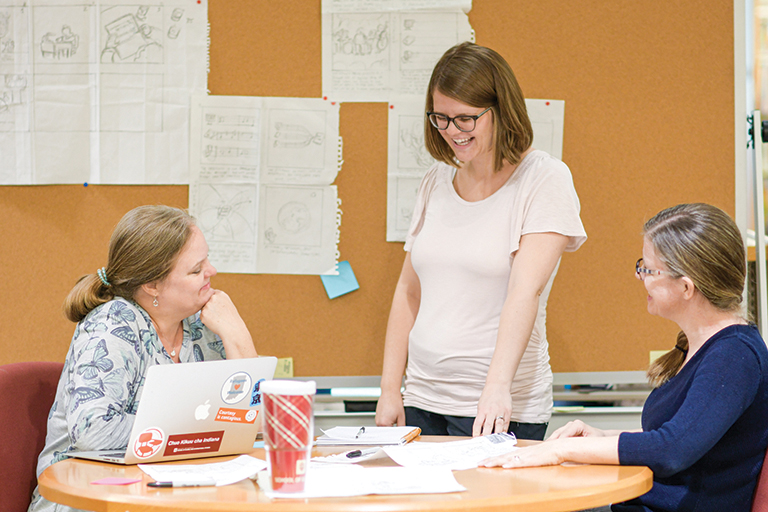 Three women are discussing work at a table where a laptop is open. On the wall are notes pinned to a board and many more notes are scattered on the table. 