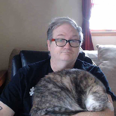 A middle aged man with glasses and a highly contented sleeping striped cat on his lap.