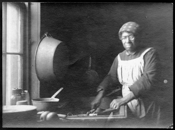 A black and white image shows a women in an kitchen wearing an apron.