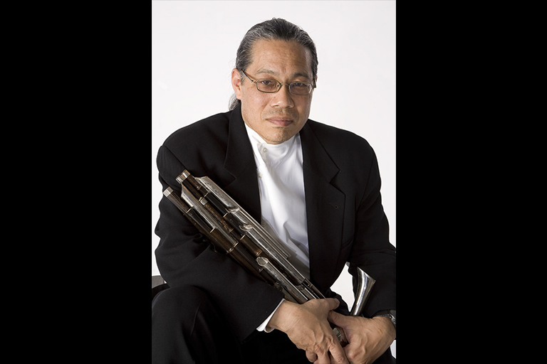 A musician in a black suit and white shirt poses for a formal portrait holding his wind instrument.