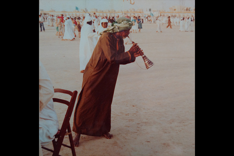 A man in a brown robe plays a horn-type instrument in a large and busy outdoor meeting place.
