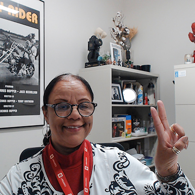 Middle-aged Black woman wearing an IU Libraries lanyard and flashing the peace sign. A vintage promotional poster for the Peter Fonda film "Easy Rider" is behind her to her left. To her right is a curio cabinet with knick-knacks.