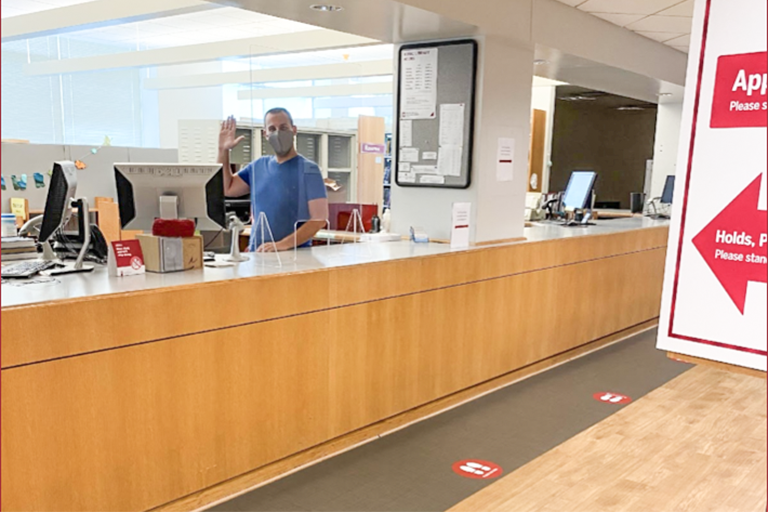 Man in protective mask and blue shirt waving from a library checkout desk. There are hand sanitizer warnings and "Stand Here" stickers on the floor. 