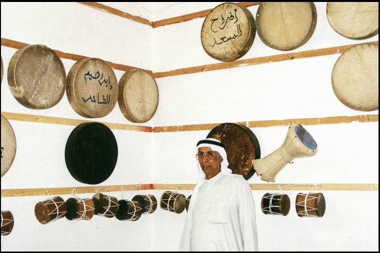 A man stands in front of a white wall on which hang a number of various hand drums.