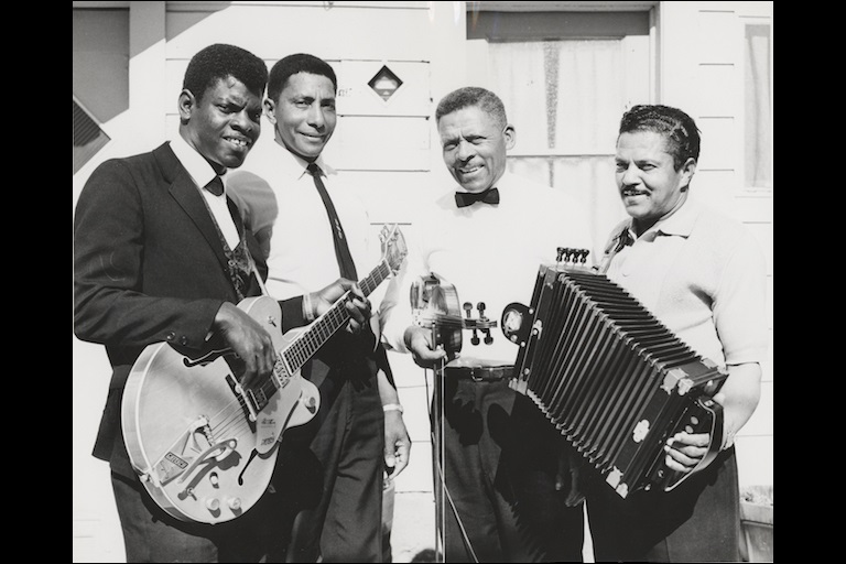 Four nicely dressed men stand together outside a home around a microphone and holding an electric guitar, fiddle, and accordian.