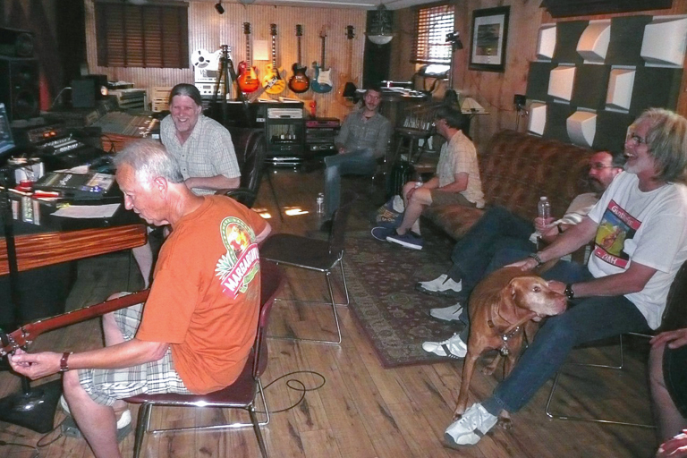 Several people sit on chairs around a recording studio, talking with each other and laughing. 