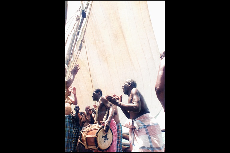 Men sing and clap and another plays a drum. A large sail is above them.