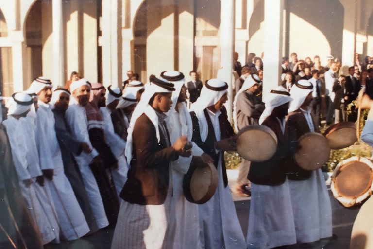 Men in robes and playing drums walk in rows in a procession past spectators under an arched colonnade.