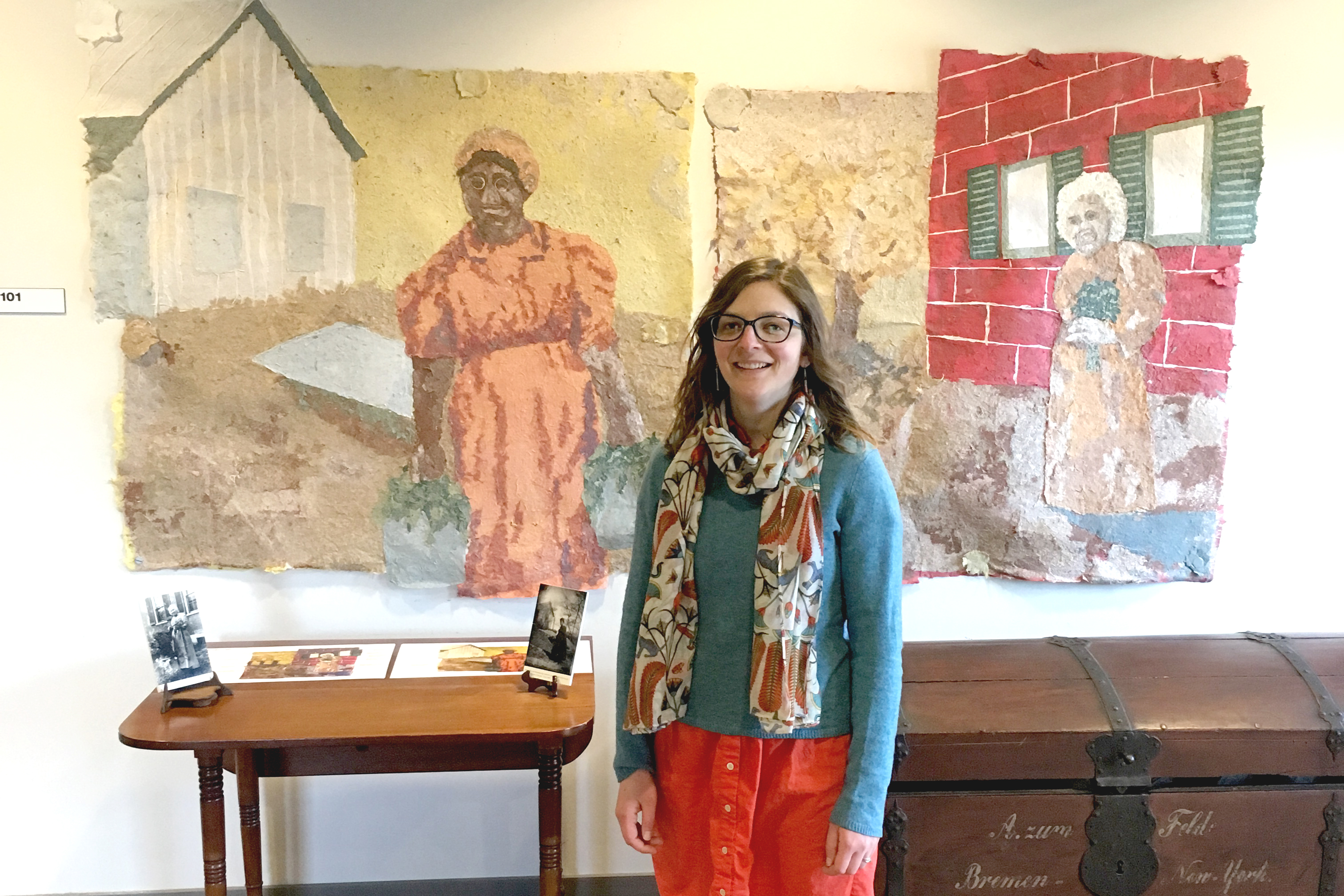 A woman, Laura Post, stands in front of her murals depicting two Wylie House historical figures.