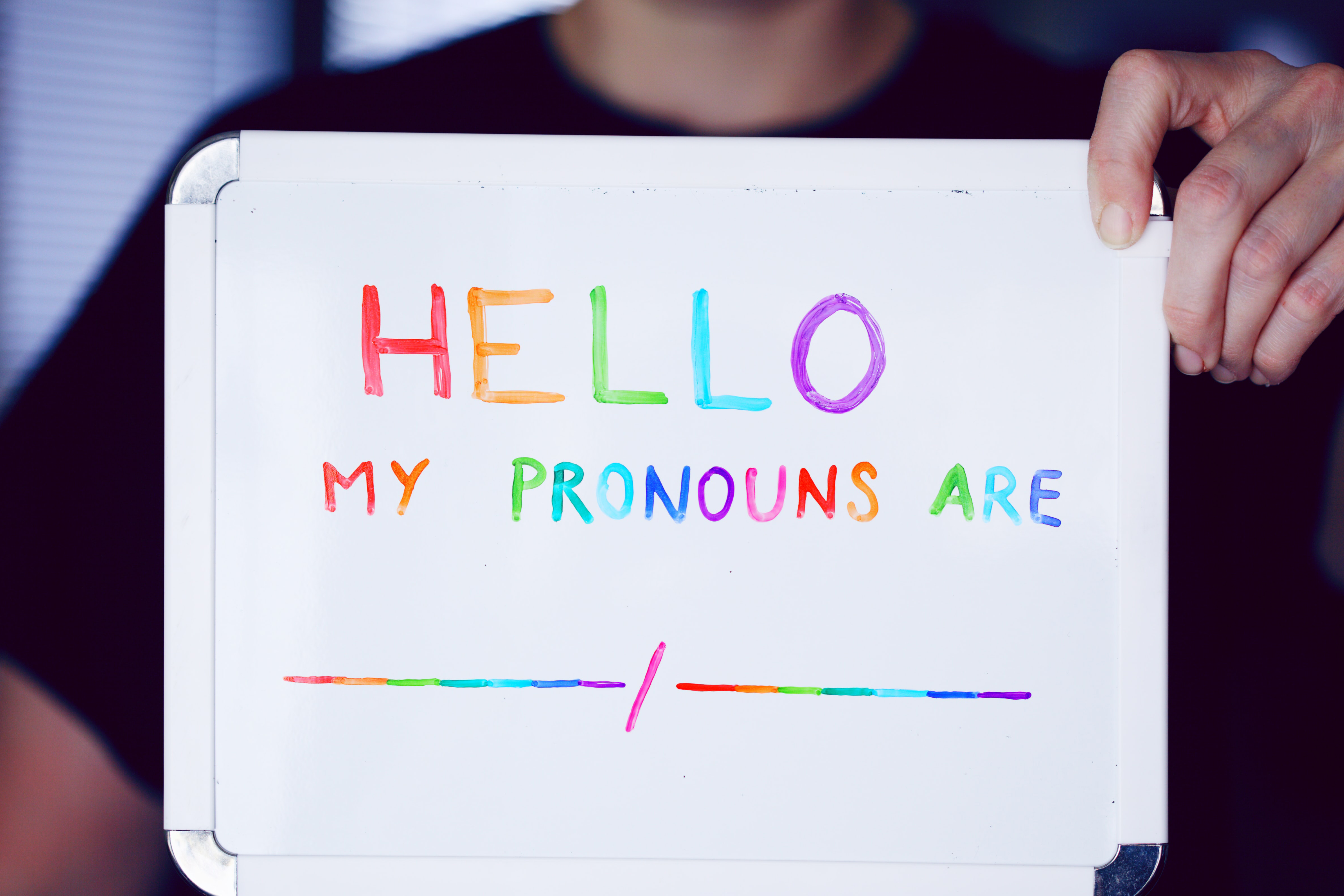 A white person holds up a whiteboard with the words "hello my pronouns are" and two blanks with a slash so one could write them in. The words are written in rainbow colors.