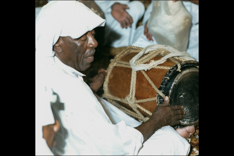 Close side profile of a man playing a drum and singing while sitting on a rug.