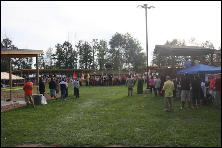 The view of a grassy entrance with a large dancing area in the background in which a group of dancers in regalia stand in a circle and are watched by standing spectators.