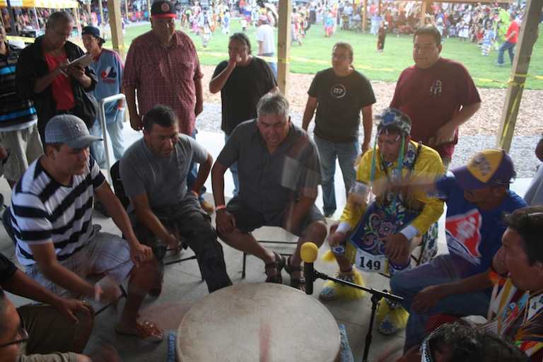Men with drum sticks sit around and bang on a large drum and sing together.