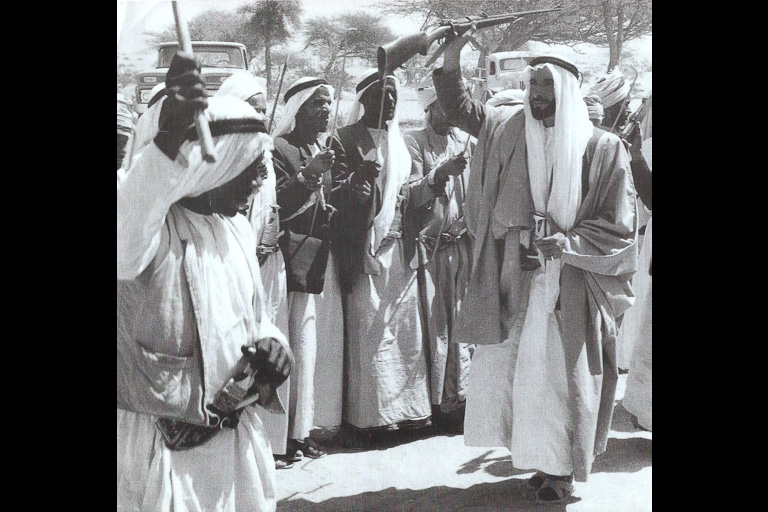 Several men stand looking at another man who holds a rifle in the air.