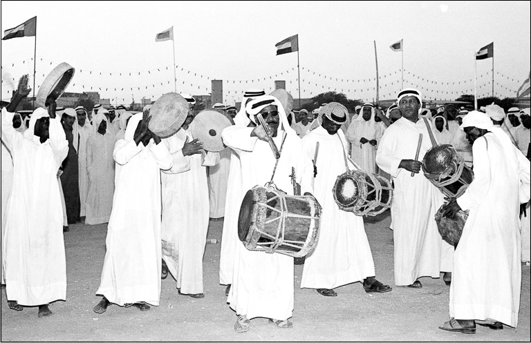 Several men stand outside in a group playing drums while many other men watch and flags fly behind them.