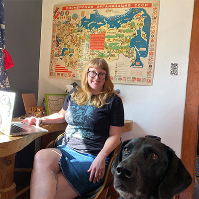 Submitted photo of a redheaded woman with thick-rimmed black glasses working in a colorful home office, with an attentive black hound dog directly in the foreground.