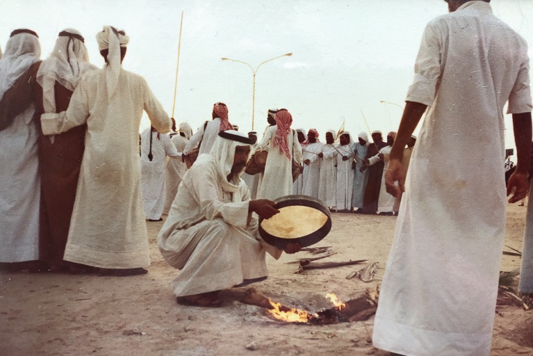 A man holding a hand drum crouches down by an open-air fire while several other men stand nearby.