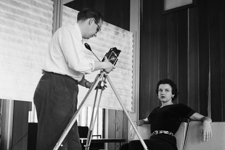 Black and white photograph of Jack Welpott taking a photo of a woman sitting on a sofa