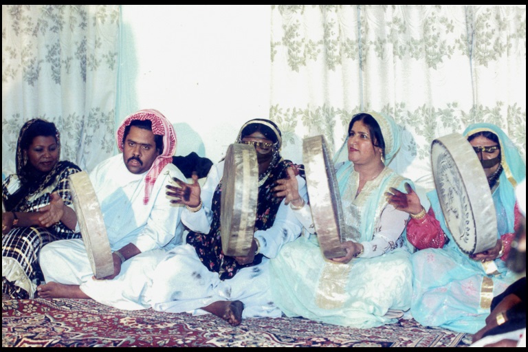 Four women and one man sit on a red rug in front of white and green drapes and play  hand drums.