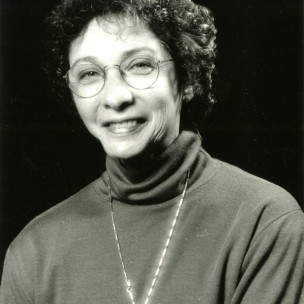 Esther Thelen smiles at the camera, wearing a turtleneck sweater, wire framed glasses, and a long oval necklace.