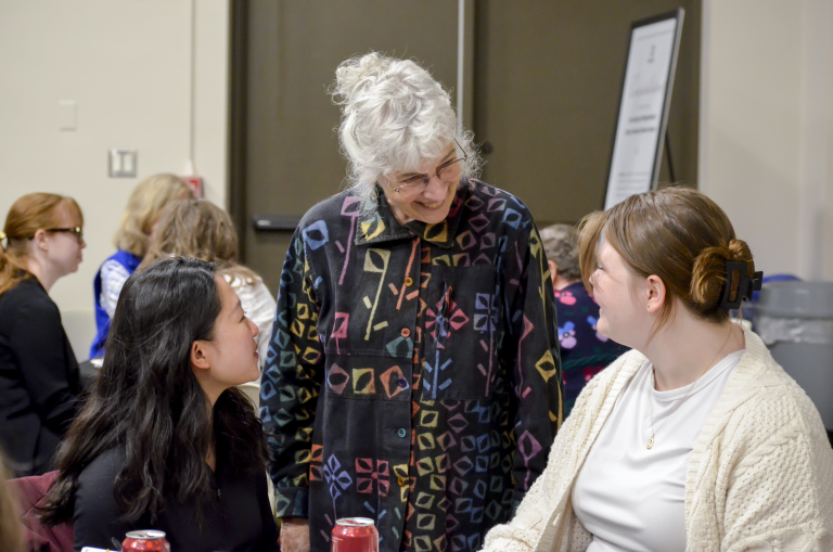 Lilly and Christina are seated at a table. Their faces are turned toward a grey-haired woman who is standing, looking down at them. Jo Burgess is the woman. She has on a jacket with brightly colored squiggles on a black background.