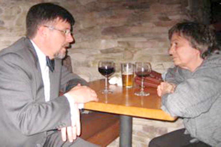 Two people lean toward each other to talk over a table with wine glasses sitting on it. The man wears a suit.