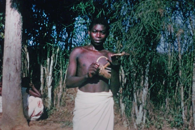 A boy stands in a jungle clearing holding a small musical instrument.