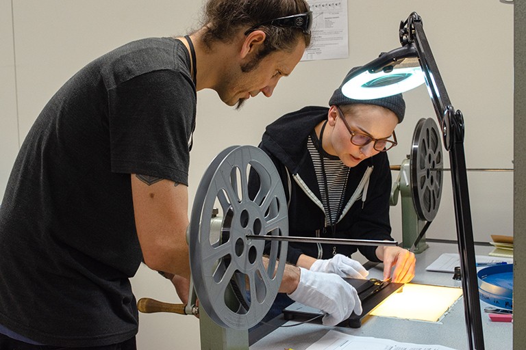 A man and a women work together at a table. Film and film reels are in the foreground.