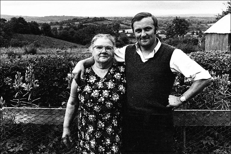 A young man stands with his arm around an older woman in rolling countryside.