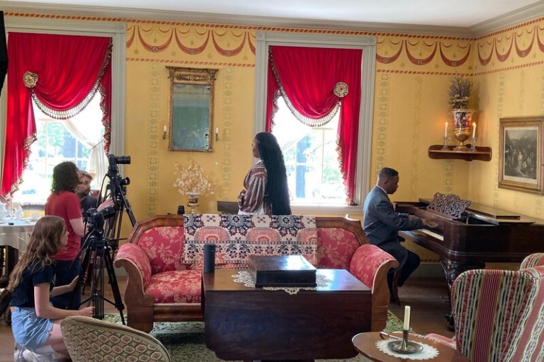 Creators of the "Stargazing" video filming in the Wylie House living room. A woman and a man perform one of the songs, wearing period costumes; several crew members cluster around a video camera.