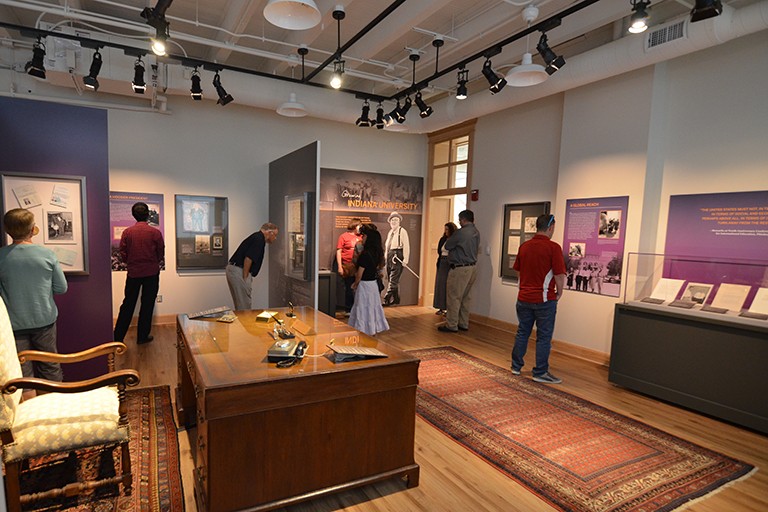 A large room set up gallery-style, with images on the walls and a large desk in the middle of the floor, and people viewing the items.